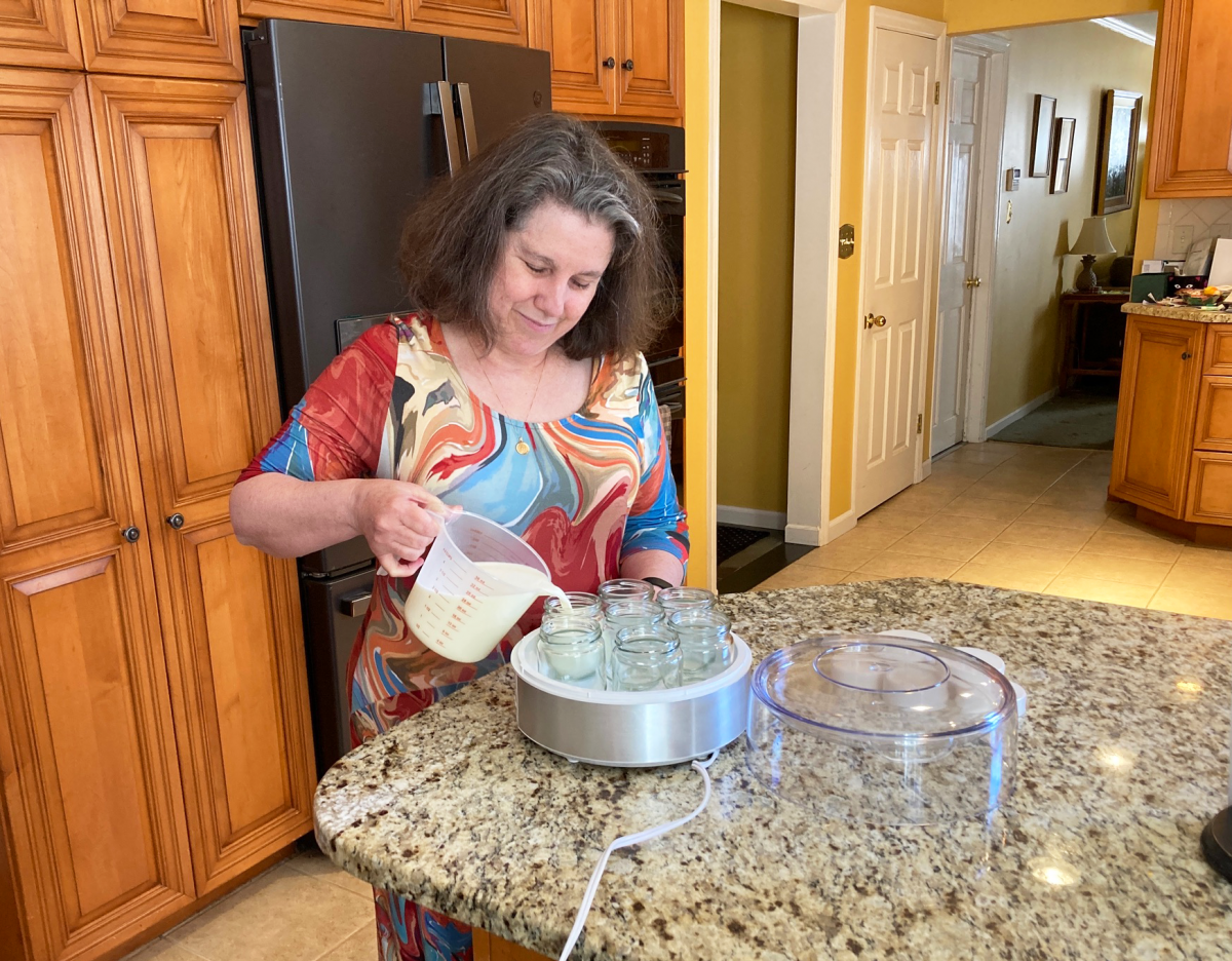 Susan Woodhouse smiles as she stands in her kitchen at home pouring liquid into jars in her yogurt maker.