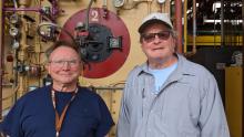 Coworkers Marshall Pysher and Dave Novogratz smile as they stand in front of Boiler #2 in the Lehigh Boiler House