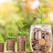 a row of coins, each successively taller from left to right, sits on a table. At the far right of the row stands a jar filled with coins representing the value of saving. Each stack of coins and the jar also have small plants growing from them, reinforcing the concept of the power of saving over time.